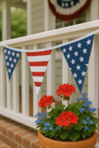 Front porch of a traditional white home decorated for the 4th of July with patriotic bunting on the railing, an American flag, red and white flowers in terracotta pots, and a navy front door with a seasonal wreath.